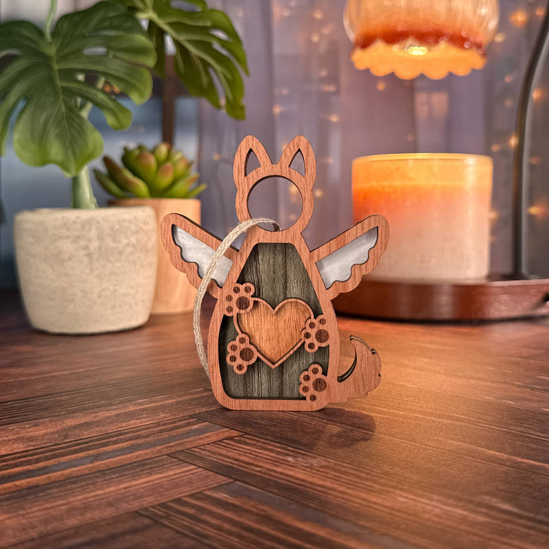 The Forged Flare® Forever By My Side Dog Angel Ornament, with wings and a heart, sits on a wooden surface—an ideal gift for dog lovers. Potted plants, a lit candle, string lights, and a glowing lamp add cozy ambiance in the background.