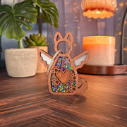 The Forged Flare® Forever By My Side Dog Angel Ornament, a wooden dog-shaped piece with angel wings and a heart cutout filled with glitter, stands on a table among potted plants and string lights—perfect for any dog lover.