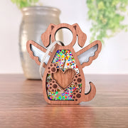 The Forged Flare® Forever In My Heart Dog Angel Ornament, featuring angel wings, a heart cutout, and colorful confetti details, stands on wood—an ideal gift for dog lovers. A blurred vase and green foliage appear in the background.