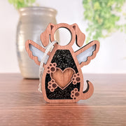 The Forged Flare® Forever In My Heart Dog Angel Ornament features angel wings, a heart and paw print design, and a string—perfect as a dog lover gift or memorial—shown on wood with a blurred vase and greenery in the background.