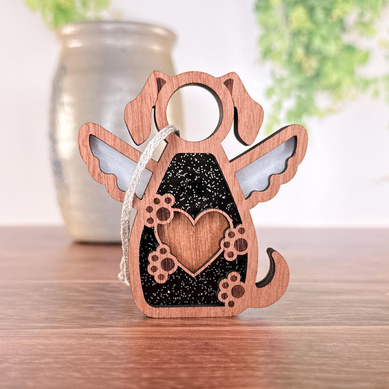 The Forged Flare® Forever In My Heart Dog Angel Ornament features angel wings, a heart and paw print design, and a string—perfect as a dog lover gift or memorial—shown on wood with a blurred vase and greenery in the background.