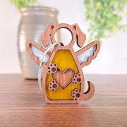 The Forged Flare® Forever In My Heart Dog Angel Ornament features a dog shape with angel wings, a yellow stained glass heart with paw prints, and a gray string. Perfect as a dog memorial or thoughtful gift for dog lovers. Blurred vase and greenery in background.