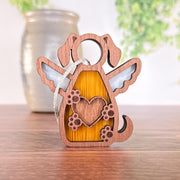 The Forged Flare® Forever In My Heart Dog Angel Ornament, featuring angel wings, a heart detail, and a hanging ribbon, rests on a wooden surface with a blurred vase and green foliage in the background.