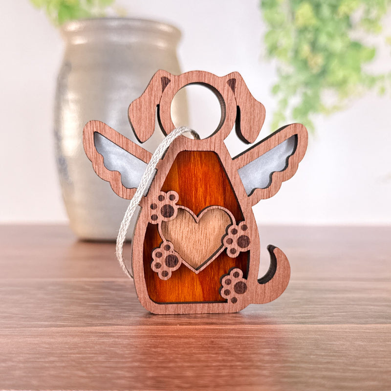 The Forged Flare® Forever In My Heart Dog Angel Ornament, a wooden angel-shaped dog memorial with a heart and paw prints, rests on wood with a blurred vase and greenery in the background.