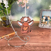 The Forged Flare® Forever In My Heart Dog Angel Ornament, with angel wings and a heart cutout, hangs on a silver stand—an ideal gift for dog lovers—beside a pen, vase of flowers, and photo frame on a wooden table.