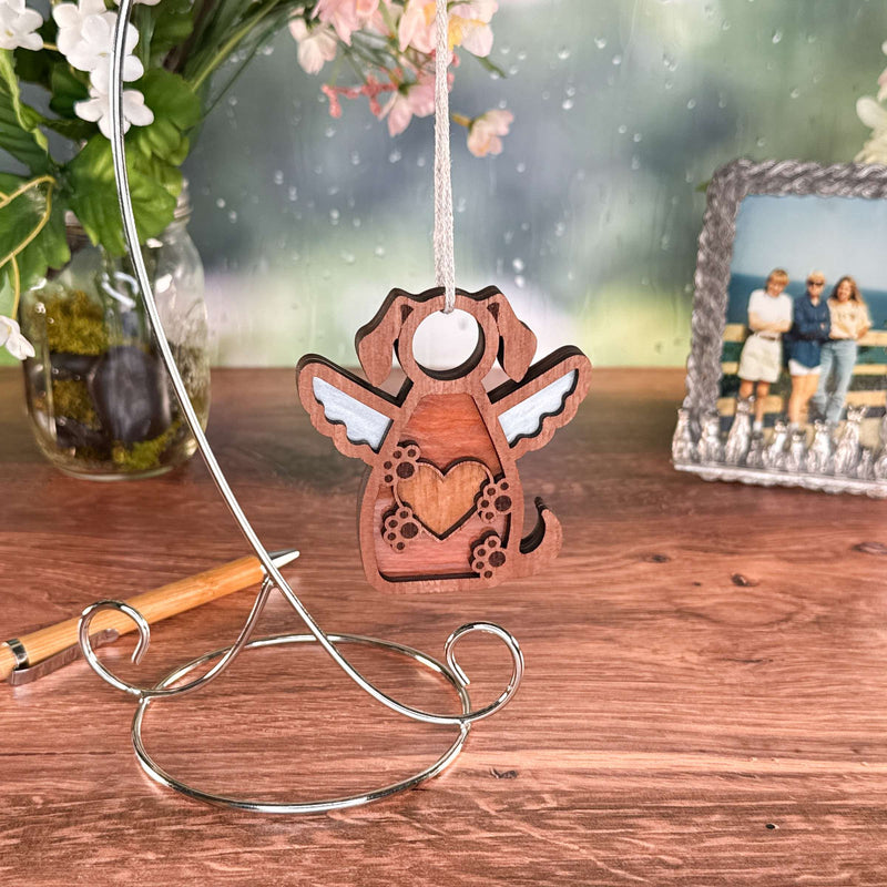 The Forged Flare® Forever In My Heart Dog Angel Ornament, with angel wings and a heart cutout, hangs on a silver stand—an ideal gift for dog lovers—beside a pen, vase of flowers, and photo frame on a wooden table.