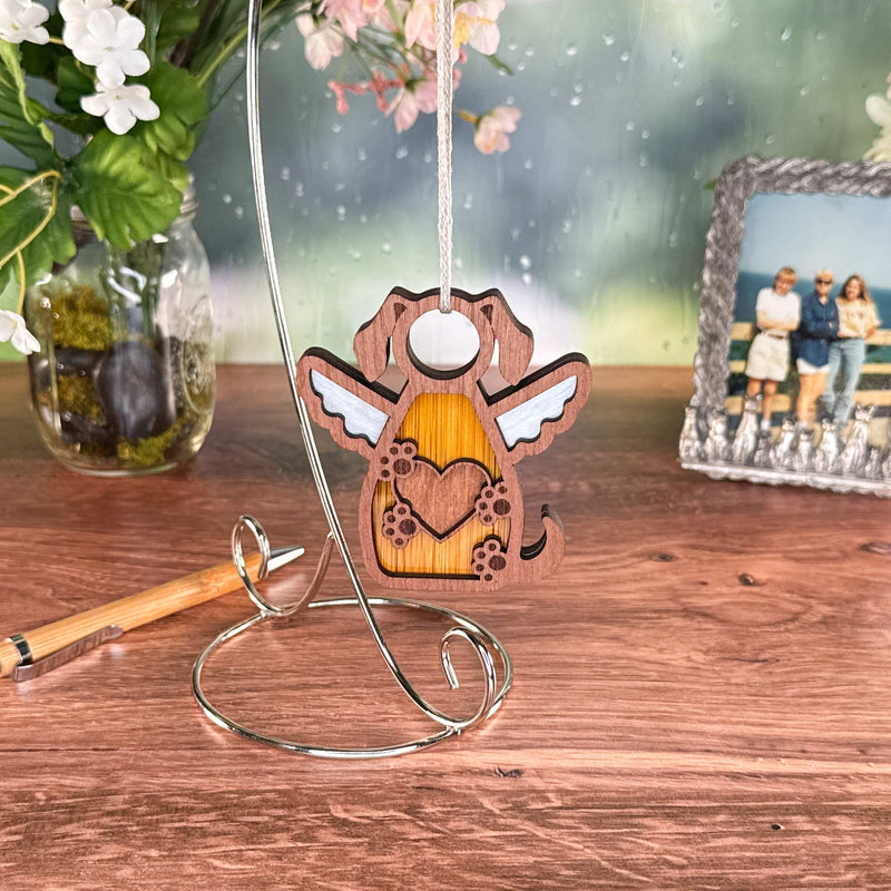 The Forged Flare® Forever In My Heart Dog Angel Ornament, featuring wings and a heart design, hangs on a curved metal stand atop wood—an ideal dog lover gift. A pen, floral vase, and framed family photo complete the background scene.