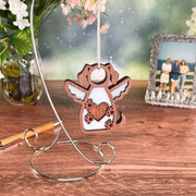 The Forged Flare® Forever In My Heart Dog Angel Ornament, shaped like a winged dog with a heart and hanging from a stand, is the perfect gift for dog lovers. It’s displayed on a wooden table with flowers, a framed photo, and a pen in the background.