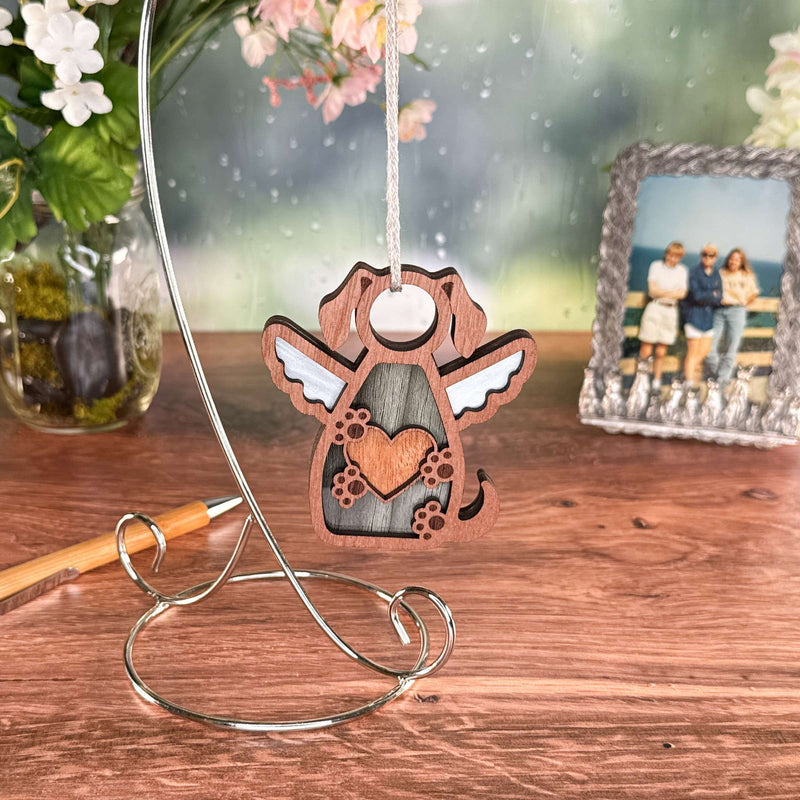The Forever In My Heart Dog Angel Ornament by Forged Flare®—featuring dog ears, wings, and a heart—hangs from a stand on a wooden table with potted flowers, a pen, and photo in the background. Ideal as a gift or memorial for dog lovers.
