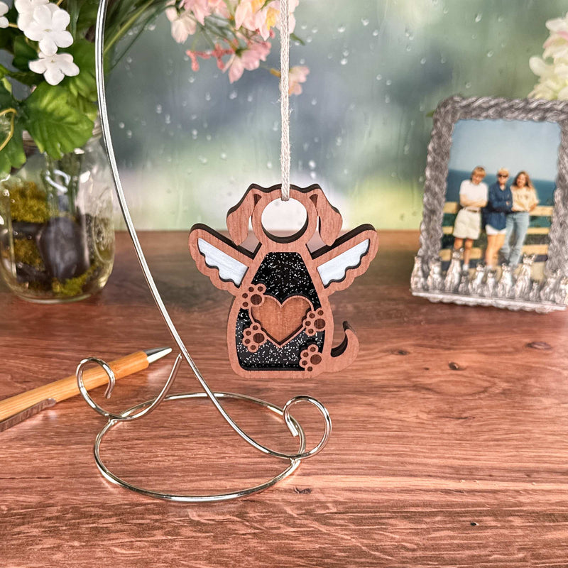 The Forever In My Heart Dog Angel Ornament by Forged Flare® features a winged fantasy dog with a heart design, hanging from a metal stand on a wooden table—an ideal gift for dog lovers. Flowers, a pen, and a framed photo sit behind it.