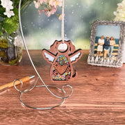 The Forged Flare® Forever In My Heart Dog Angel Ornament, a glittery wooden keepsake with a heart cutout, hangs from a silver stand on a table beside flowers, a pen, and a beach photo—perfect for dog lovers or as a memorial gift.