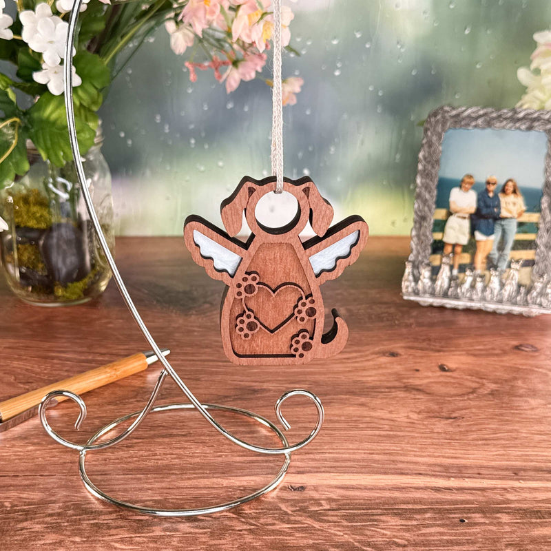 The Forged Flare® Forever In My Heart Dog Angel Ornament with wings and a heart hangs from a stand on a wooden table, with flowers and a photo in the background—making it a meaningful gift for dog lovers.