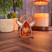 The Forged Flare® Forever In My Heart Dog Angel Ornament—a wooden dog figure with angel wings and a heart cutout—rests on wood, surrounded by a candle, potted succulent, leafy plant, and glowing string lights. Perfect as a memorial or dog lover gift.