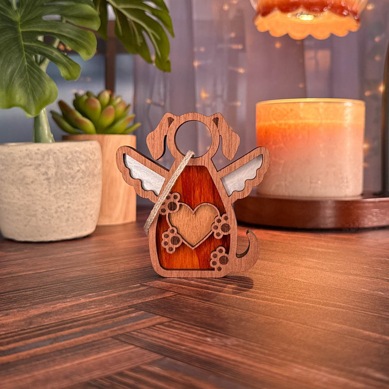 The Forged Flare® Forever In My Heart Dog Angel Ornament—a wooden dog figure with angel wings and a heart cutout—rests on wood, surrounded by a candle, potted succulent, leafy plant, and glowing string lights. Perfect as a memorial or dog lover gift.