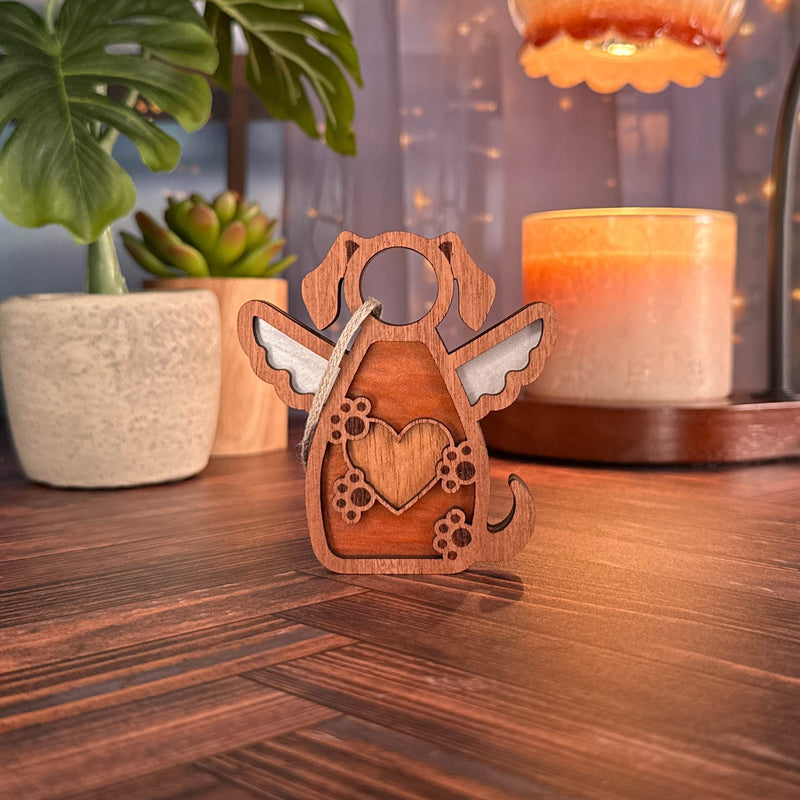 The Forged Flare® Forever In My Heart Dog Angel Ornament, featuring a heart and wings design, sits on a wooden table with plants, a candle, and string lights—a perfect dog lover gift or memorial keepsake.