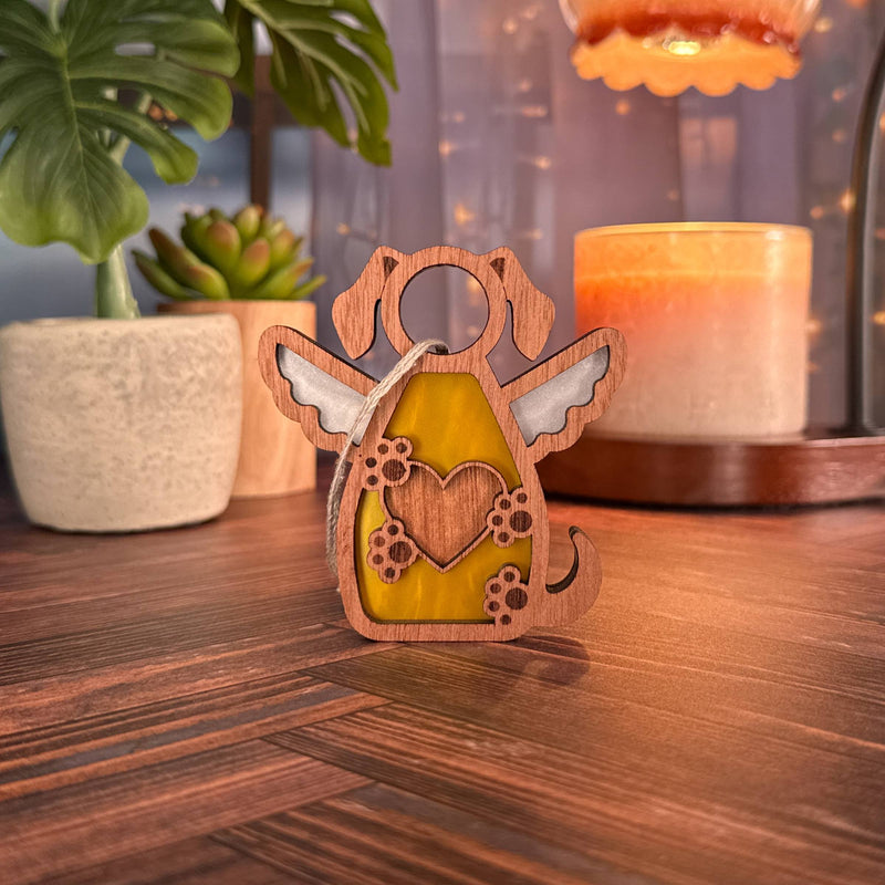 The Forged Flare® Forever In My Heart Dog Angel Ornament with stained glass wings and paw prints stands on a wooden table, surrounded by potted plants and candles—a touching dog memorial or heartfelt gift for dog lovers.