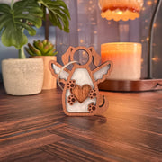 The Forged Flare® Forever In My Heart Dog Angel Ornament, featuring a heart and paw prints, stands on a table—perfect as a dog lover gift or memorial. A candle, plant, and soft lights behind it create a warm indoor scene.