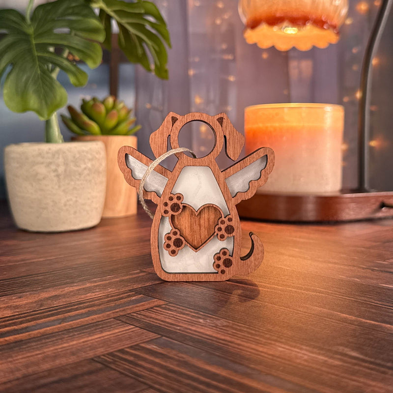 The Forged Flare® Forever In My Heart Dog Angel Ornament, featuring a heart and paw prints, stands on a table—perfect as a dog lover gift or memorial. A candle, plant, and soft lights behind it create a warm indoor scene.