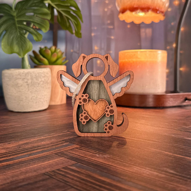 The Forged Flare® Forever In My Heart Dog Angel Ornament—a small wooden dog figurine with wings, halo, and heart—sits on a wooden surface. A touching memorial for dogs and perfect gift for dog lovers. Houseplants and soft lights blur behind it.