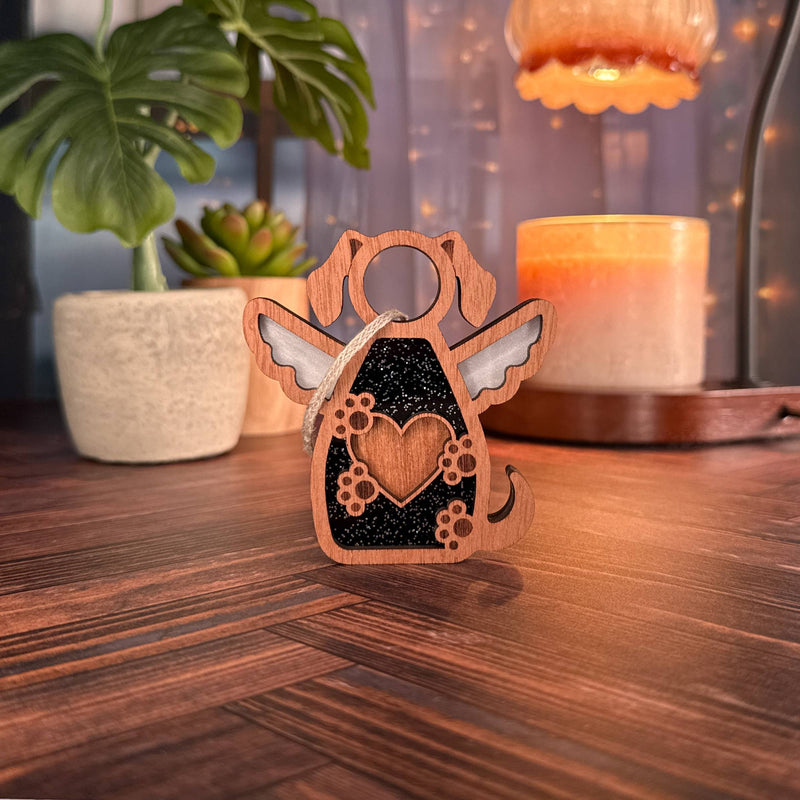 The Forged Flare® Forever In My Heart Dog Angel Ornament, a wooden angel with wings and a heart center, sits on a table surrounded by candles, a plant, and string lights—an ideal gift for any dog lover.