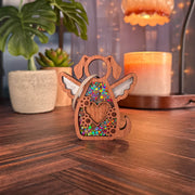 The Forged Flare® Forever In My Heart Dog Angel Ornament, shaped like a winged pup with a heart cutout and glitter accents, sits on a wooden table—an ideal gift for dog lovers. Soft lighting and fairy lights glow in the blurred background.
