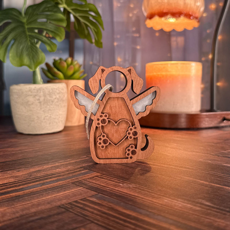 The Forged Flare® Forever In My Heart Dog Angel Ornament—a wooden dog with wings and heart—rests on a cozy surface among potted plants and candlelight, making a perfect personalized gift for any dog lover.