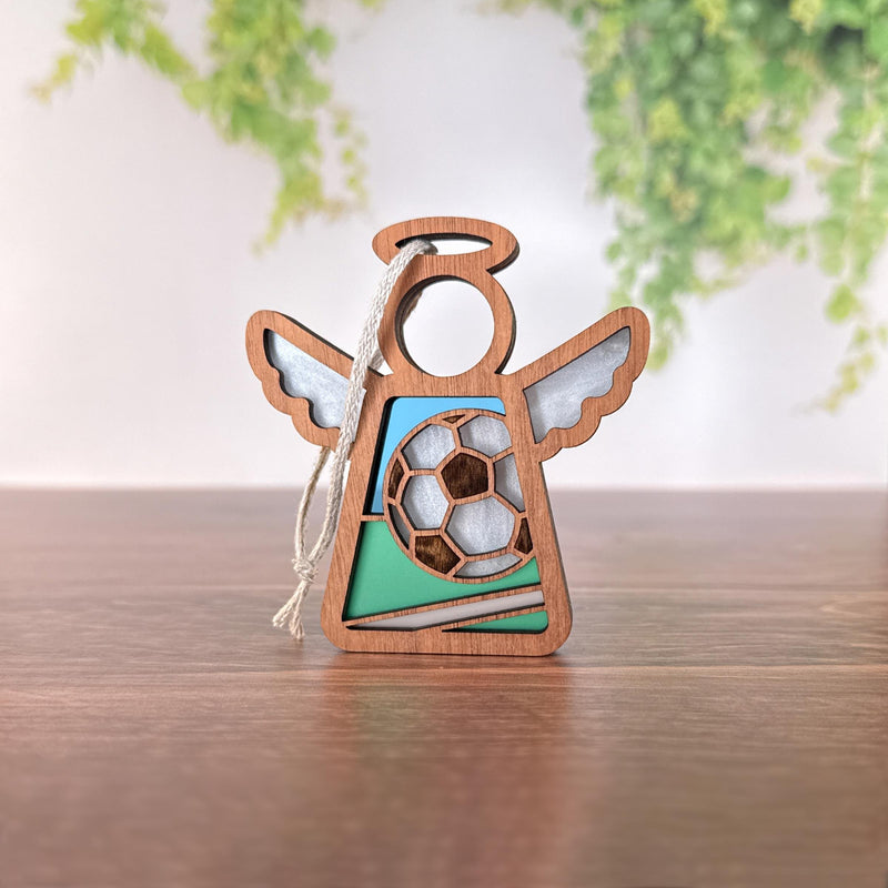 The Soccer Ornament | 3.5" (H) Angel Figurine by Forged Flare® features a wooden angel with a soccer ball cutout—an ideal Mother's Angels® soccer team Christmas gift, displayed on wood with leafy greenery in the background.