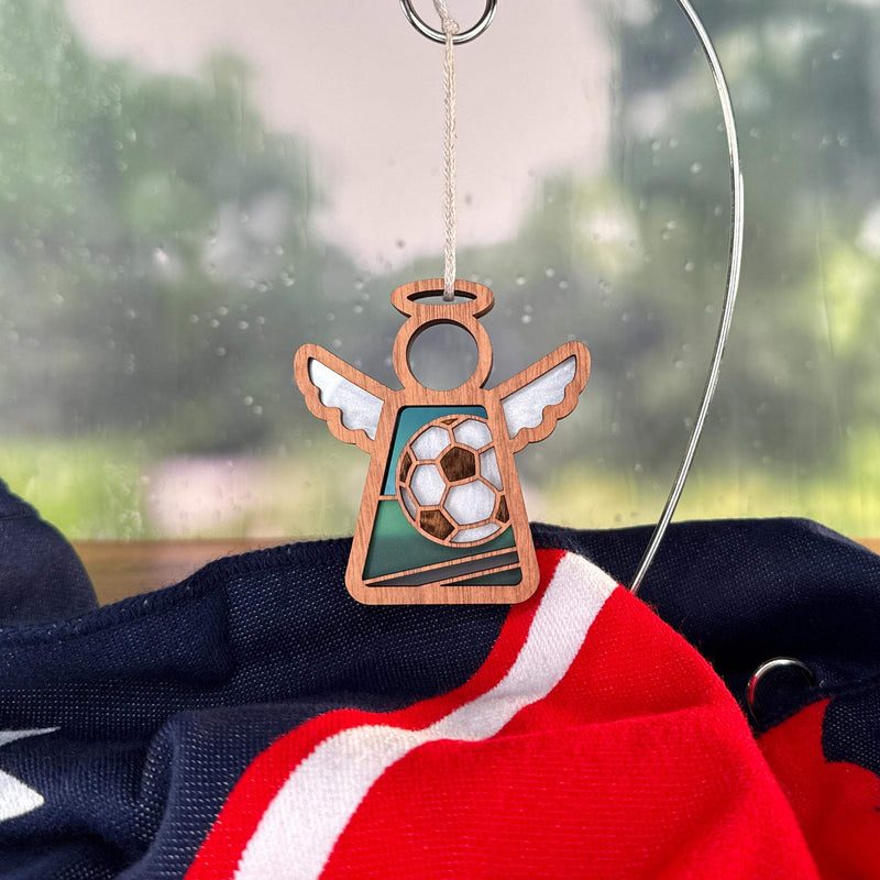 The Forged Flare® Soccer Ornament | 3.5" (H) Angel Figurine from Mother's Angels® hangs on a metal hook before a rainy window, above a dark blue, red, and white cloth—an ideal soccer coach appreciation gift.