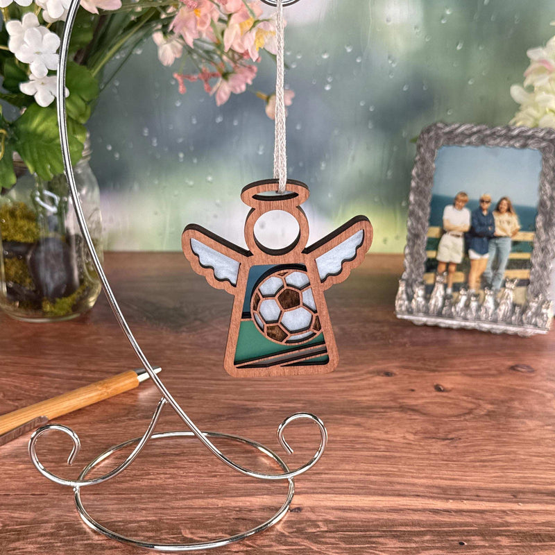 The Forged Flare® Soccer Ornament | 3.5" (H) Mother's Angels® angel figurine stands on a wooden table near flowers and a framed photo—an soccer themed gift for a soccer-loving fan.