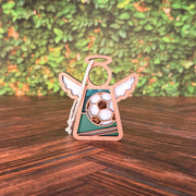 The Forged Flare® Soccer Ornament— a 3.5" Mother's Angels® angel figurine featuring a white soccer ball design—stands on a wooden surface, making a charming unique soccer Christmas gift against a backdrop of blurred greenery.