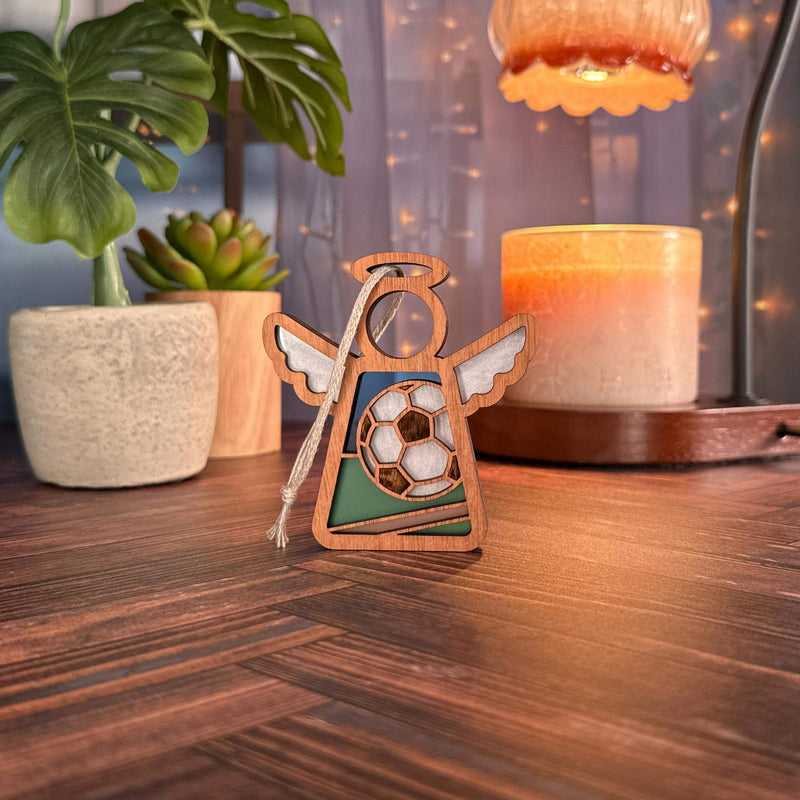 The Soccer Ornament | 3.5" (H) Angel Figurine by Forged Flare® stands on a wooden table with plants, a candle, lamp, and string lights behind it—creating a cozy scene and making it a perfect soccer coach appreciation gift.