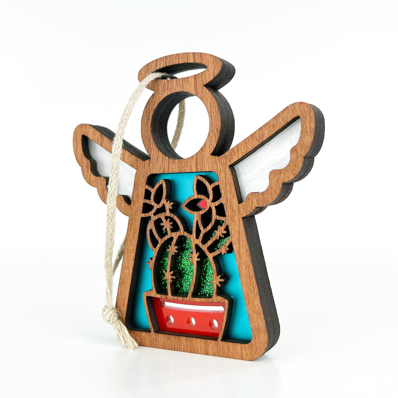 The Desert Cactus Ornament by Forged Flare® is a 3.5" wooden angel figurine with a halo and cut-out cacti design in a red pot against a blue background. Includes white hanging string—perfect for Southwest decor. Part of Mother's Angels®.