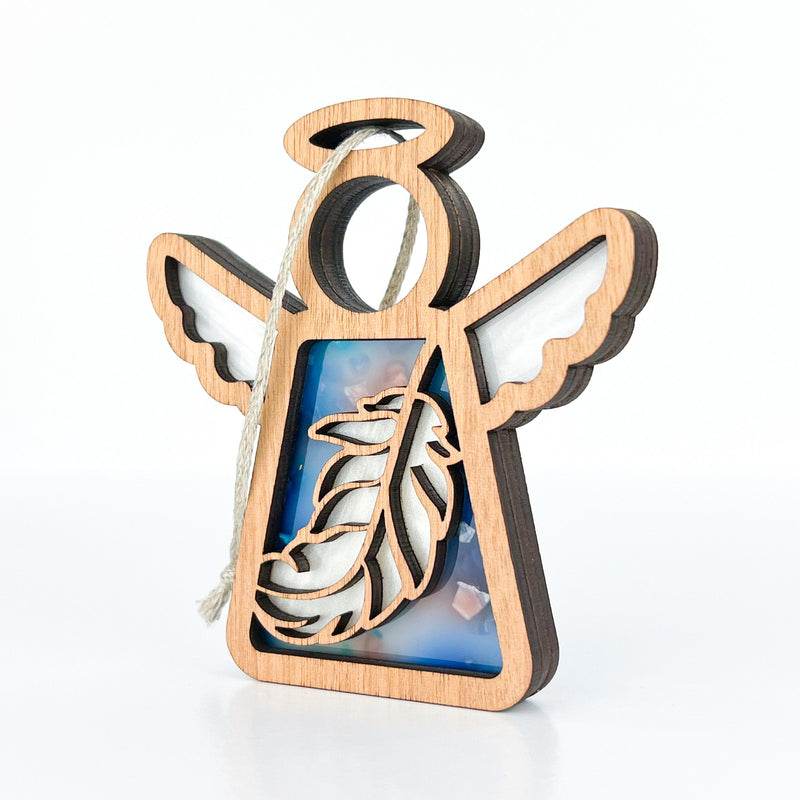 The Mother's Angels® Feather Ornament by Forged Flare® is a 3.5" wooden angel figurine with cut-out wings, feather design, a blue and white resin background, and string loop—ideal as a mindfulness gift or unique Christmas ornament.