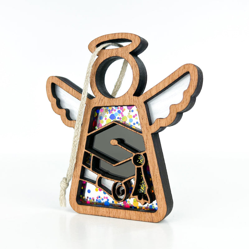 The Graduation Ornament from Forged Flare®'s Mother's Angels® collection is a 3.5" wooden angel figurine adorned with a halo and wings. Designed with a black graduation cap, diploma, and colorful confetti within its body, this cut-out ornament comes with an attached string for easy hanging. It's perfect for adding to your angel figurines collection or as decor for graduation parties.