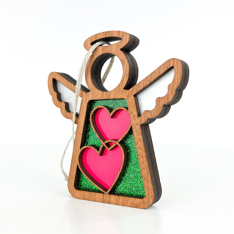 The Forged Flare® Limited Edition Interlocking Hearts Ornament - Series 8 is a 3.5" wooden angel with white wings, a green glitter background, and two pink hearts, complete with string for hanging as a festive Mother's Angels® decoration.