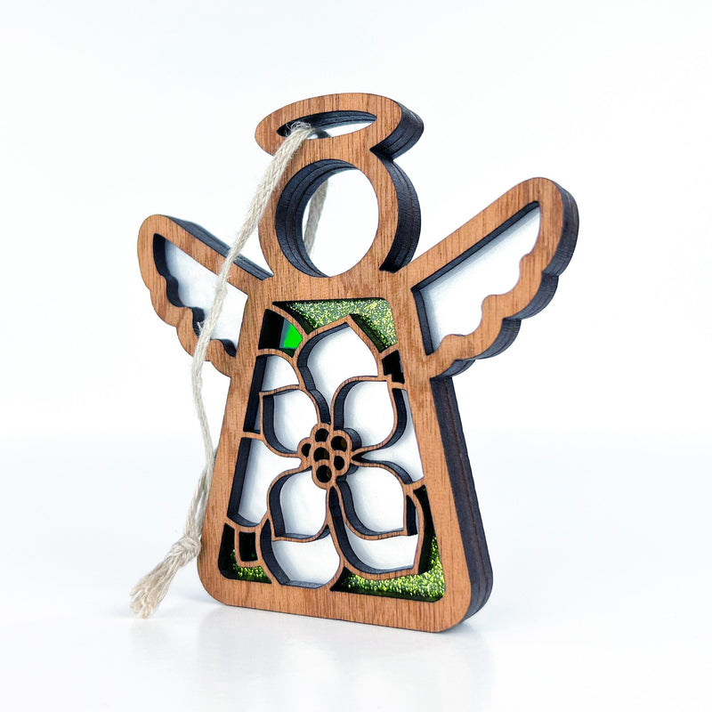The Magnolia Ornament by Forged Flare® is a 3.5" angel figurine from the Mother's Angels® collection. This wooden angel features open wings, a halo, and a cutout flower design that reveals green glitter behind it. Ideal for Christmas tree decoration, it comes with a loop of white string for easy hanging.