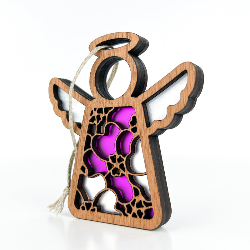 A delicate 3.5" angel figurine from Forged Flare's® Mother's Angels® collection, named the Primrose Ornament, features an intricate floral cutout design against a vibrant purple background and showcases the February birth flower, pink primrose. With a halo and outstretched wings, this wooden angel hangs gracefully from a loop of twine and is displayed on a plain white surface.