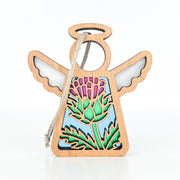 The Forged Flare® Scotch Thistle Ornament is a 3.5" Mother's Angels® figurine featuring a wooden angel with a cut-out thistle flower design, symbolizing strength and resilience, plus a hanging string and light blue backdrop.