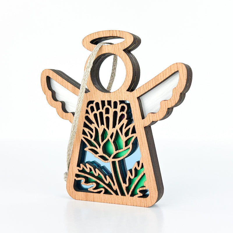The Forged Flare® Scotch Thistle Ornament is a 3.5" Mother's Angels® figurine with cut-out wings, a halo, and green-blue stained glass thistle symbolizing strength and grace. It hangs easily—perfect as a unique Christmas ornament.