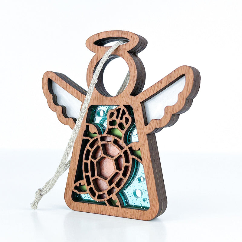 The Forged Flare® Sea Turtle with Imperfections Ornament is a 3.5" Mother's Angels® wooden angel figurine with painted white wings, a halo, turtle cutout, green and blue accents, and a hanging string—perfect for Christmas or coastal decor.