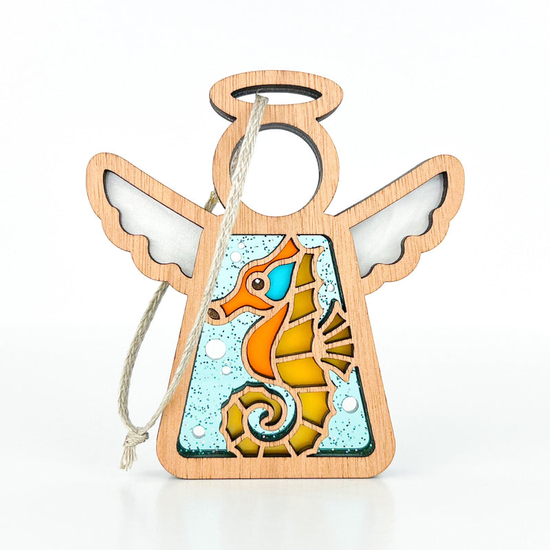 The Forged Flare® Seahorse Ornament is a 3.5" wooden Mother's Angels® figurine with white wings, a halo, and a vibrant stained-glass seahorse design—ideal as a unique Christmas tree ornament or beach house decor. Includes hanging string; background is white.