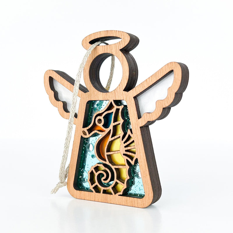 The Forged Flare® Seahorse Ornament is a 3.5" wooden angel figurine from Mother's Angels®, with outstretched wings, seahorse and ocean cut-outs, layered colorful details, and a white hanging string—perfect for Christmas trees or beach house décor.