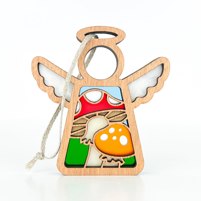 The Forged Flare® Mother's Angels® Toadstool Mushroom Ornament is a 3.5" wooden angel figurine with white wings, holding a red and yellow stained-glass style toadstool. Includes string for hanging as a Christmas tree ornament. Plain white background.