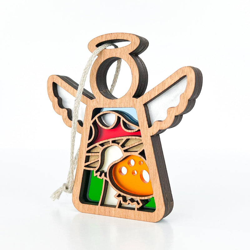 The Forged Flare® Mother's Angels® Toadstool Mushroom Ornament is a 3.5" angel-shaped wooden figurine with cutout wings and halo, colorful toadstools and greenery inside, plus a white hanging string. Plain white background.