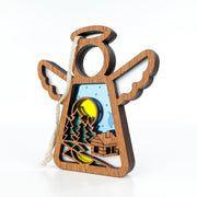The Forged Flare® Warm Welcome Ornament with Imperfections is a 3.5" handcrafted Mother's Angels® wooden angel featuring a cutout winter scene—trees, house, and yellow moon or sun against a blue sky with snowflakes—ready to hang.