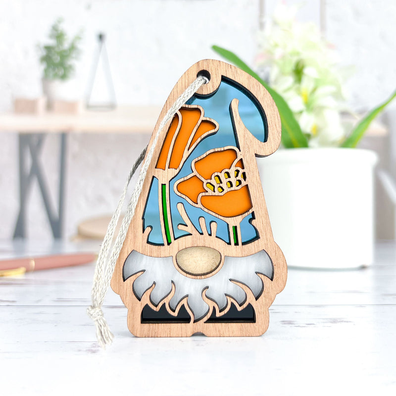 The Forged Flare® California Poppy Ornament (3.75" H) from the Nature's Gnomes™ collection sits on a white surface, featuring orange poppies and a blue background. A potted plant and blurred home decor are in the background.