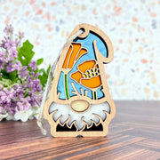 The Forged Flare® Nature’s Gnomes™ California Poppy Ornament, a 3.75" garden gnome with a stained glass-style orange poppy and blue background, sits on a table with purple and white flowers behind it.