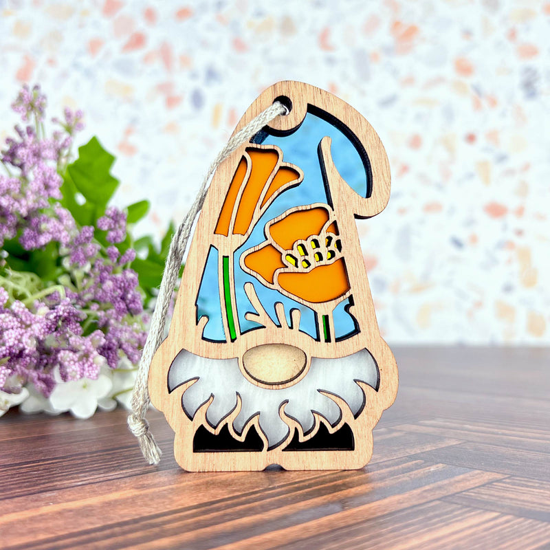 The Forged Flare® Nature’s Gnomes™ California Poppy Ornament, a 3.75" garden gnome with a stained glass-style orange poppy and blue background, sits on a table with purple and white flowers behind it.