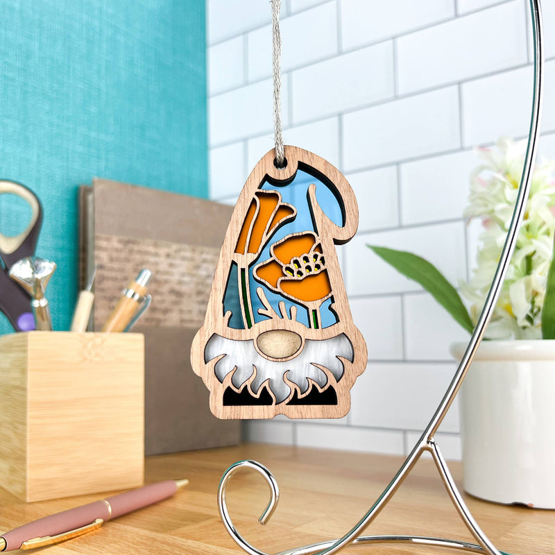 The California Poppy Ornament, a 3.75" wooden garden gnome from Forged Flare’s Nature’s Gnomes™ collection, features a blue hat and orange flower design and hangs in a bright, modern workspace with office supplies and flowers visible.