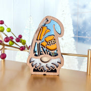 The California Poppy Ornament by Forged Flare®—a 3.75" Nature's Gnomes™ garden gnome with a white beard, yellow flowers, and sky-blue background—stands on a table amid blurred frosted trees and decorative berries.